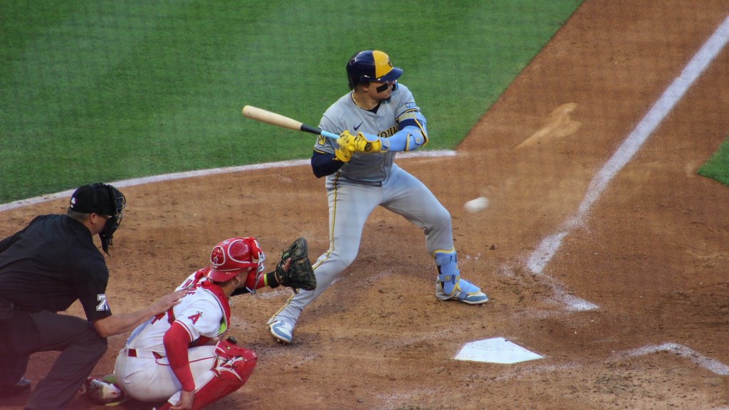Brewers catcher William Contreras, in a gray road uniform, squares up in the batter's box as a pitch comes in, with the Angels catcher and the umpire crouched behind the plate.