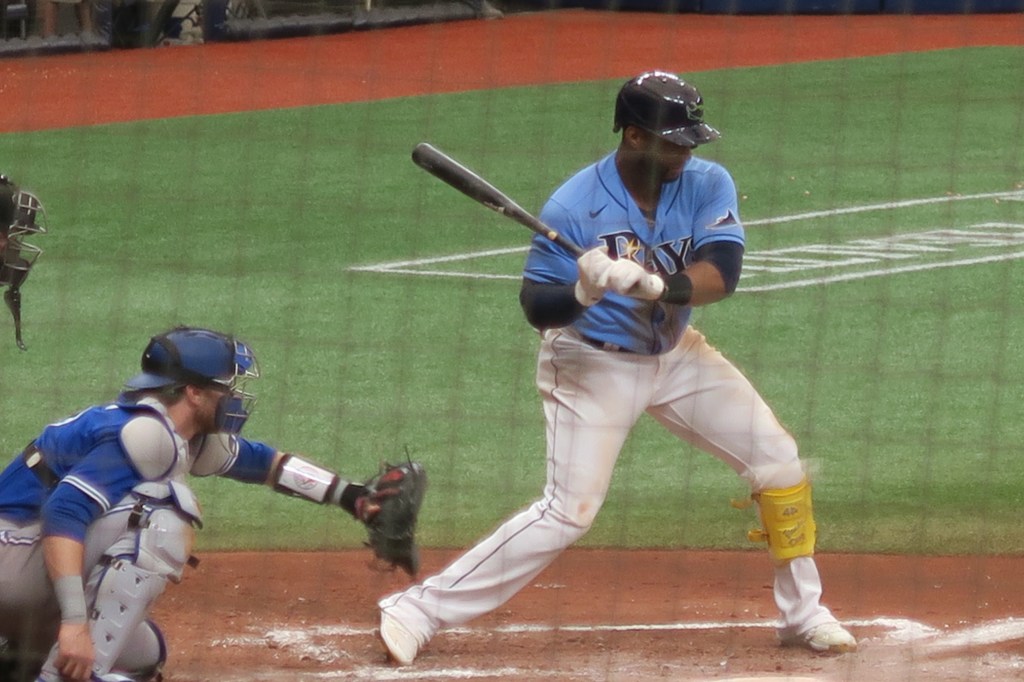 Tampa Bay Rays hitter Yandy Díaz in a light-blue Rays uniform at the plate, watching a pitch, with the catcher crouched behind him.
